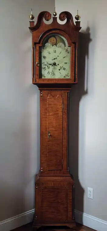 Tall case clock with a figured wood case and a painted dial, standing in a hallway
